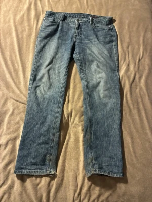 Blå raka jeans med slitna detaljer - Ett par klassiska blå jeans med rak passform och slitna detaljer vid bensluten. Jeansen har fem fickor och synliga sömmar. Perfekta för en avslappnad och trendig look.