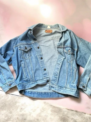 Klassisk blå Levi's jeansjacka - Säljer en klassisk blå jeansjacka från Levi's med raka sömmar, två bröstfickor med lock och knapp samt knappstängning framtill. Jackan har en avslappnad passform och tidlös design som passar till det mesta. Perfekt för dig som gillar vintage och streetstyle.