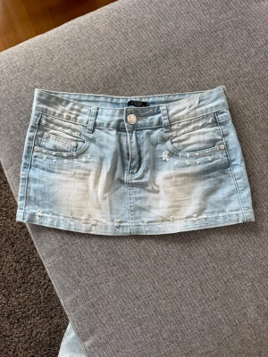 Ljusblå jeanskjol med slutningar  - Ljusblå jeanskjol med slitna detaljer och dekorativa nitar vid fickorna. Kjolen har klassisk femficksdesign, bälteshällor och knappstängning framtill. Perfekt för en avslappnad och cool stil.