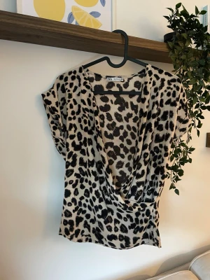 Leopardmönstrad omlottopp från Zara - Snygg omlottopp från Zara med leopardmönster i svart och beige. Toppen har djup v-ringning och korta ärmar, perfekt för att sticka ut lite extra. Tillverkad i mjukt och lätt material som känns skönt mot huden.