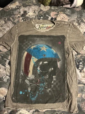 Amplified t-shirt med dödskalletryck - Cool beige t-shirt från Amplified med ett stort tryck av en dödskalle med hjälm i blått, vitt och rött framtill. T-shirten har rund hals och korta ärmar. Materialet känns mjukt och lätt, perfekt för en avslappnad stil.