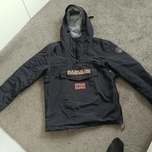 Svart vindjacka från Napapijri - Snygg svart vindjacka från Napapijri med stor huva och dragkedja vid sidan. Jackan har en stor ficka framtill med norsk flagga och Napapijri-logga, samt ett märke på ärmen. Insidan är fodrad med grått tyg. Perfekt för blåsiga dagar.