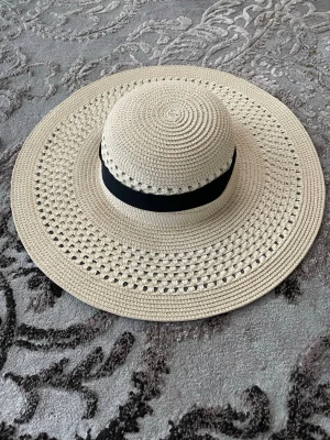 Beige solhatt med svart band - Snygg beige solhatt med bred skärm och dekorativt svart band runt kullen. Hatten är virkad i ett luftigt material som ger en cool och avslappnad look. Perfekt för att skydda ansiktet mot solen och lyfta din outfit på stranden eller i stan.