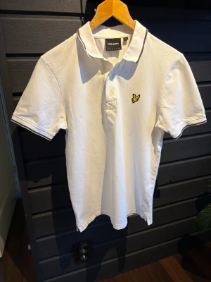 Vit pikétröja från Lyle & Scott - Klassisk vit pikétröja från Lyle & Scott med korta ärmar och gul logga på bröstet. Tröjan har krage med knappar och diskreta mörka ränder längs kragen. Perfekt till en avslappnad och stilren look.