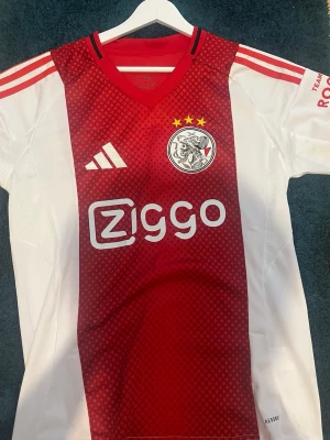 Ajax 25/26 home - Ajax officiella matchtröja från Adidas i rött och vitt med klubbmärke, tre stjärnor och Ziggo-logga på bröstet. Tröjan har inte varit använd någon gång och är helt ny. Men tröjan är stor i sin storlek