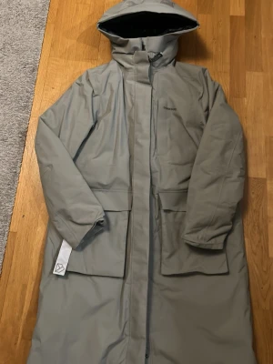 Ljusgrå regnjacka från Didriksons - Snygg ljusgrå regnjacka från Didriksons med huva och långa ärmar. Jackan har två stora fickor framtill och dragkedja med vindslå. Materialet är vattenavvisande och perfekt för regniga dagar. Enkel, stilren design med Didriksons-logga på bröstet.