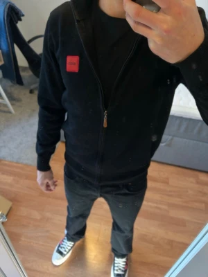 Svart zip hoodie från Hugo - Snygg svart hoodie från Hugo med dragkedja framtill och röd logotyp-patch på bröstet. Klassisk modell med huva och ribbade muddar. Tillverkad i mjukt bomullsmaterial som känns skönt mot huden. Perfekt till jeans eller joggers för en clean look.