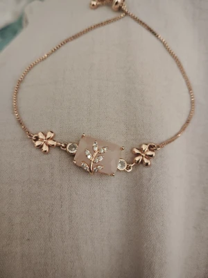 Roséguldfärgat armband med blommor - Snyggt armband i guld med två blommor och en rektangulär sten i mitten, dekorerad med gnistrande stenar i ett bladliknande mönster. Kedjan är tunn och justerbar för perfekt passform. Perfekt accessoar för att lyfta din outfit.