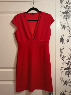 Röd kortklänning med v-ringning - Snygg röd kortklänning med djup v-ringning och markerad midja. Klänningen har korta ärmar och en enkel, stilren design som passar perfekt till många tillfällen. Tillverkad i ett mjukt och stretchigt material för en bekväm passform.