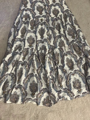 Vit långkjol med bohemiskt mönster - Långkjol i vitt med bohemiskt, paisley-inspirerat mönster i blått och brunt. Kjolen har volanger och en luftig passform som ger en avslappnad vibe. Perfekt för dig som gillar en unik och stilren look.