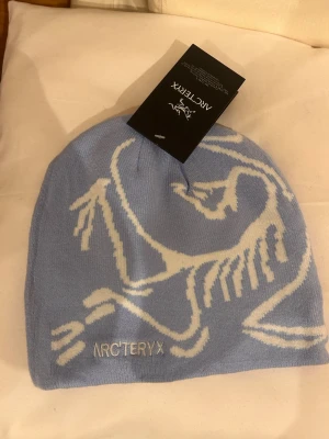 Ljusblå mössa från Arc'teryx - Snygg ljusblå mössa från Arc'teryx med vitt fossil-motiv och broderad logga framtill. Tillverkad i mjukt, stickat material som sitter skönt på huvudet. Perfekt för dig som vill ha en stilren och sportig accessoar till vintern.