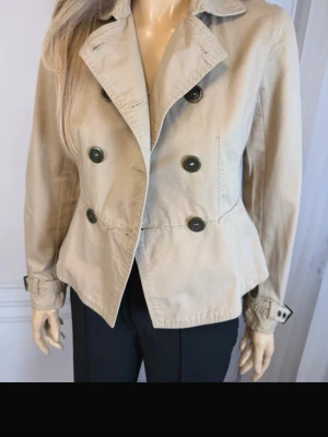 Kort beige trenchcoat med knappar - Stilren kort trenchcoat i beige med dubbelknäppning och stora svarta knappar. Jackan har klassisk krage, markerad midja och justerbara spännen vid ärmsluten. Perfekt för dig som gillar en tidlös och enkel look. Materialet ser ut att vara bomull eller bomullsblandning.