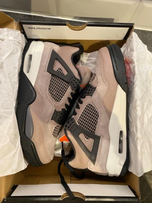 Air Jordan 4 Taupe Haze sneakers - Säljer ett par Air Jordan 4 Taupe Haze sneakers med beige mocka, svarta och grå detaljer samt meshpaneler. Skorna har en vit och svart sula med synlig Air-enhet och orange inslag vid plösen. Klassisk siluett och cool design.