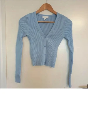 Ljusblå ribbad kofta med v-ringning - Snygg ljusblå ribbad kofta med v-ringning och långa ärmar. Koftan har knäppning framtill med matchande knappar och är tillverkad i ett mjukt, stretchigt material. Perfekt att slänga över en topp för en chill look.