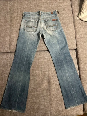 Ljusblå bootcut jeans från 7 For All Mankind - Säljer ett par ljusblå bootcut jeans från 7 For All Mankind med snygg slitning och klassiska kontrastsömmar på bakfickorna. Jeansen har fem fickor och är tillverkade i mjuk denim med en avslappnad vibe. Negativt är att en av öglorna vid midjan lossnat, annars är skicket på jeansen hur bra som helst.