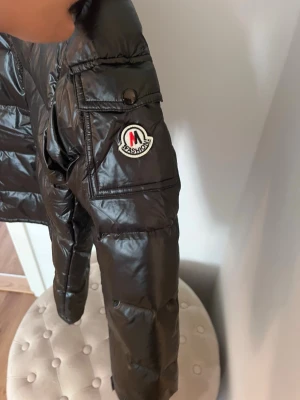 Svart dunjacka från fashion  - Snygg svart dunjacka från fashion med glansig finish och vadderad design. Jackan har en ficka med knapp på ärmen och en tydlig logotyp-patch. Utrustad med huva och breda kanaler för extra värme, perfekt för kalla dagar.