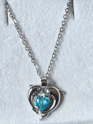 Delfinhalsband med Ljusblå Zirkon - Silverton - Nytt - Ett charmigt och romantiskt halsband med en delfindesign som omsluter en vacker, hjärtformad syntetisk zirkon i en klar ljusblå/ turkos nyans. Den glittrande stenen ger smycket en lyxig känsla och en härlig färgklick.Perfekt som en fin present eller för att addera en lekfull och glänsande detalj till din egen outfit. En tidlös design för alla som älskar havets mest älskade djur!  Detaljer: Design: Delfin med hjärtformad sten  Färg: Silverton med ljusblå/turkos sten  Sten: Syntetisk zirkon (Kubis