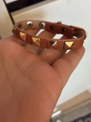 Valentino Brunt läderarmband med nitar - Oäkta Snyggt brunt armband i läder med guldiga pyramidformade nitar runt om. Justerbar storlek med tryckknappar för perfekt passform. Perfekt accessoar för att ge din outfit en edgy touch.