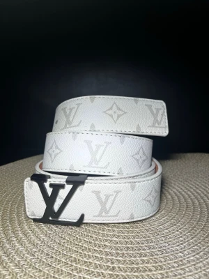 Vitt Louis Vuitton bälte med monogram - Snyggt vitt bälte från Louis Vuitton med klassiskt monogrammönster och stor LV-logga i metall som spänne. Tillverkat i skinn och har exklusiv känsla, perfekt för att lyfta vilken outfit som helst.