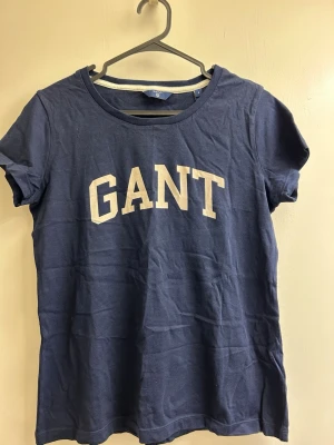 Marinblå GANT t-shirt i bomull - Klassisk marinblå t-shirt från GANT med vit logotyp framtill. T-shirten har rund halsringning och korta ärmar. Tillverkad i mjuk bomull för en skön och avslappnad stil.