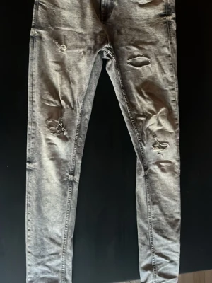 Grå slitna skinny jeans från FSBN - Säljer ett par grå skinny jeans från FSBN med slitningar och hål på benen för en trendig look. Jeansen har en tvättad finish och smal passform som sitter snyggt. Perfekta för dig som gillar streetstyle och vill ha ett par bekväma men coola jeans.