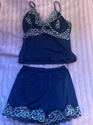 Mörkblå spetsigt pyjamasset - Säljer ett mörkblått pyjamasset med linne och shorts. Linnet har tunna axelband och spetsdetaljer i vitt över bysten och under bysten. Shortsen har matchande vit spets längs nederkanten. Setet är gjort i ett mjukt, följsamt material som känns lyxigt mot huden.
