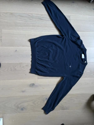 Mörkblå tröja från Gant - Klassisk mörkblå tröja från Gant med rund halsringning och diskret logga broderad på bröstet. Tröjan har långa ärmar och ribbade muddar vid ärmslut och nederkant. Perfekt för en stilren och enkel look.