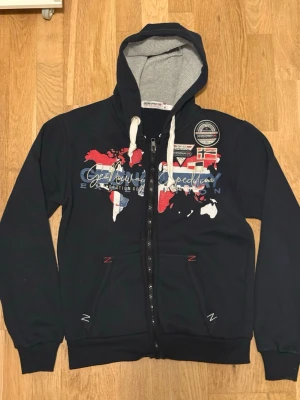 Geographical normal zip-up (mörkblå) - Snygg mörkblå hoodie från Geographical Norway med dragkedja, vit insida på huvan och tryck av världskarta och flaggor på bröstet. Fickor framtill och vita snören i huvan. Perfekt för en cool och avslappnad stil.