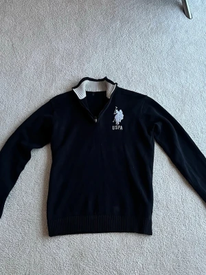 Svart USPA tröja med halv dragkedja - Svart långärmad tröja från USPA med halv dragkedja och vit logga broderad på bröstet. Kragen har en snygg vit insida med svarta ränder. Tröjan har ribbade muddar vid ärmslut och nederkant. Perfekt för en stilren och sportig look.
