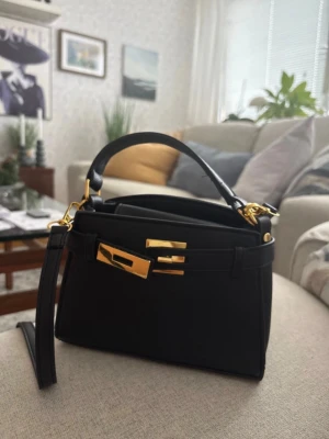 Svart minihandväska med gulddetaljer - Snygg svart minihandväska i slätt läder med guldfärgade metalldetaljer och ett kort handtag. Väskan har en stilren design med dekorativa spännen framtill och avtagbar axelrem. Perfekt accessoar för dig som gillar minimalistisk och lyxig stil.