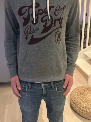 Grå Superdry hoodie - Säljer en grå hoodie från Superdry med stort vinrött tryck på bröstet och vita dragsnören i huvan. Hoodien har känguruficka framtill och ribbade muddar vid ärmslut och nederkant. Perfekt för en avslappnad streetstyle look. Den är i mycket bra skick och riktigt fet och jag är 173