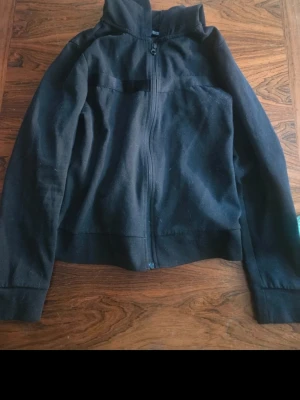 Svart hoodie med dragkedja - En enkel svart hoodie med dragkedja framtill och huva. Modellen har långa ärmar och breda muddar vid ärmslut och nederkant. Perfekt för en avslappnad och stilren look. Materialet ser ut att vara mjukt och bekvämt, troligen bomullsblandning.