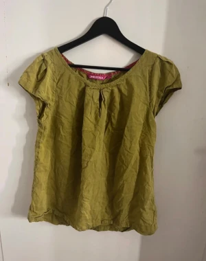 Gulaktig kortärmad blus  - En enkel och luftig gulaktig blus med rund halsringning och liten vikt detalj framtill. Blusen har korta ärmar och är tillverkad i ett lätt tyg som känns skönt mot huden. Perfekt att matcha med jeans eller kjol för en avslappnad look. Grön/gulaktig färg och skönt material. Passar alla väder! 