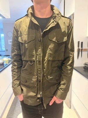 Olivgrön parkajacka med dragkedja - Snygg olivgrön parkajacka med flera stora fickor, axelklaffar och detaljer som knappstängning och dragkedja framtill. Jackan har en modern, normal passform och är tillverkad i ett lätt syntetmaterial. Perfekt för dig som gillar stilrena och funktionella plagg.