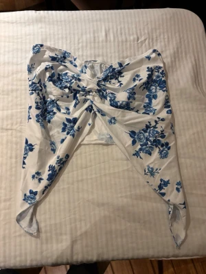 Vit och blå blommig bandeau topp - Säljer en bandeau topp i vitt med blått blommönster. Toppen har en knytning framtill som ger en cool rosettdetalj och är axelbandslös. 