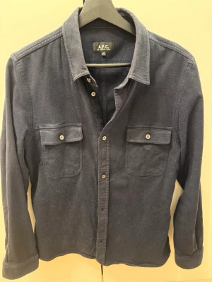 Mörkblå skjorta/overshirt från A.P.C. - Snygg mörkblå skjorta från A.P.C. i bomull med klassisk krage och två bröstfickor med lock och knappar. Skjortan har långa ärmar och knäppning framtill. Perfekt för en clean och stilren look.