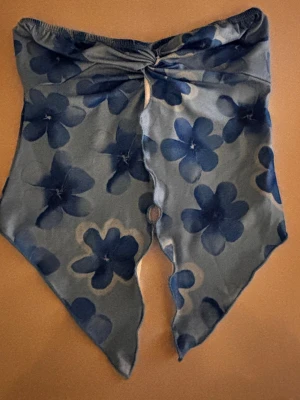 Blommig blå bandeau tubtopp - Trendig bandeau tubtopp med stora blå blommor på ljus bakgrund. Toppen har en spetsig nederkant och twistad detalj framtill. Perfekt för sommarens alla tillfällen och enkel att styla med jeans eller kjol.