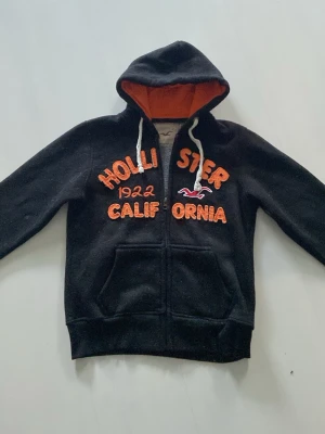 Svart Hollister hoodie med orange text - Svart hoodie från Hollister med dragkedja, orange broderad text och logga på bröstet samt vita snören i huvan. 
