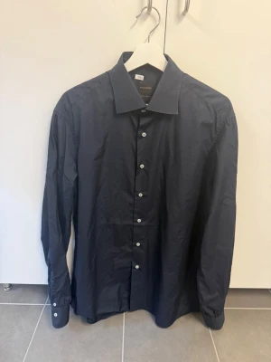 Mörkblå skjorta från Rose & Born stl M - Klassisk mörkblå skjorta från Rose & Born med knappar framtill och lång ärm. Skjortan har en stilren design och är tillverkad i bomull, perfekt för dig som gillar en clean och tidlös look.