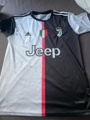 Juventus matchtröja Adidas svart/vit - Juventus officiella matchtröja från Adidas i svart och vitt med röd rand i mitten. Tryck med Jeep-logga, klubbmärke och italienska skölden på framsidan. På ryggen står det 'Ronaldo' och nummer 7. Tillverkad i lätt och ventilerande material.