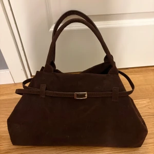 Mörkbrun mocka handväska med bälte - Snygg mörkbrun handväska i mjuk mocka med dubbla handtag och dekorativt bälte med metallspänne framtill. Väskan har en klassisk rektangulär form och passar perfekt för dig som gillar stilrena accessoarer.