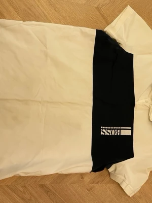 Vit och svart BOSS t-shirt - Snygg vit t-shirt från BOSS med en bred svart panel över bröstet och ett stort BOSS-tryck i vitt. Klassisk passform med korta ärmar och rund hals. Perfekt för dig som gillar stilrena och moderna plagg.