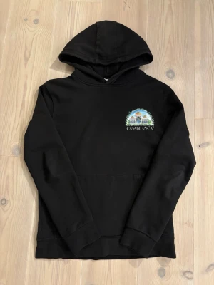  Casablanca hoodie - Svart hoodie från Casablanca med färgglad broderad grafik och text på bröstet samt stor illustration och text på ryggen. Klassisk känguruficka och huva. Perfekt för dig som gillar streetwear med unik design.