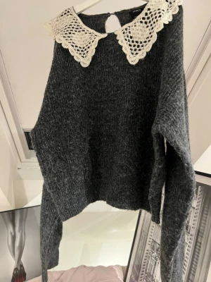 Grå stickad tröja med spetskrage - Mysig grå stickad tröja från Lindex med en stor, vit virkad spetskrage som ger en vintagekänsla. Tröjan har lång ärm och är gjord i ett fluffigt material som känns både trendigt och bekvämt. Perfekt för dig som gillar unika detaljer.