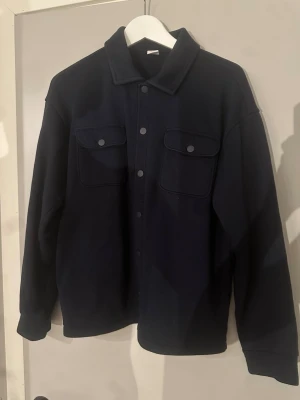 Mörkblå overshirt med fickor - Stilren mörkblå overshirt med klassisk krage och tryckknappar framtill. Två stora bröstfickor med lock och knapp ger en cool utility-look. Overshirten har raka ärmar och är gjord i ett mjukt, tjockare tyg som passar perfekt till lager-på-lager.