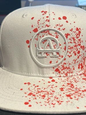 Vit keps All Cap one limited edition keps - Snygg vit keps med platt skärm och broderad logga framtill. Unik design med röda färgstänk över hela framsidan och skärmen. Märket All Cap x Chris står broderat på sidan. Perfekt för dig som vill sticka ut med din stil. Aldrig använd