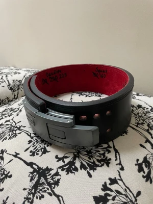 SBD lyftarbälte 13mm - Säljer ett svart lyftarbälte från SBD med röd insida och metallspänne. Bältet är tillverkat i kraftigt läder och har justerbart spänne för optimal passform vid styrkelyft och gymträning, dessvärre har jag skrivit lite gamla pbn på insidan men som kan tvättas bort👍