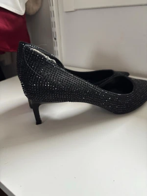 Svarta pumps med strass från Zara - Säljer ett par svarta pumps från Zara med spetsig tå och låg klack. Skorna är täckta av små svarta strasstenar som ger en glittrig look. Tillverkade i syntetmaterial och har en elegant siluett som sticker ut.