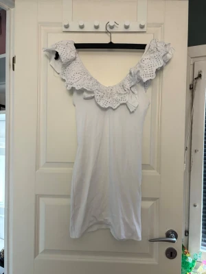 Vit klänning med volang från Zara - Supersöt vit klänning från Zara med bred volangkrage i broderad spets och ärmlös design. Klänningen är kort och har en romantisk känsla, perfekt för varma dagar. Tillverkad i mjukt material som känns skönt mot huden.