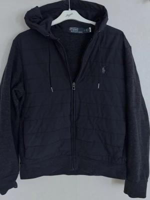 Svart cardiganjacka från Polo Ralph Lauren - Mörkgrå jacka med dragkedja från Polo Ralph Lauren. Jackan har quiltad front, mjuka ärmar och huva med dragsko. Liten broderad logga på bröstet. Materialmix med vadderad framsida och sweatshirt-tyg på ärmar och rygg.